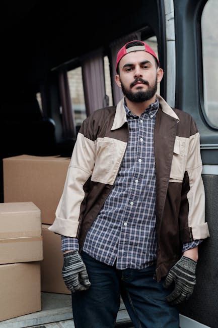 A young man with a beard, wearing a black cap and a white polo shirt, stands inside the open rear of a moving van loaded with numerous cardboard boxes of various sizes, some secured with plastic strap