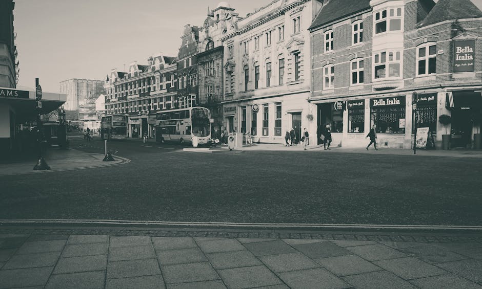 A street scene in Preston featuring a row of multi-storey Victorian-style buildings with brick facades and large windows, some with decorative cornices, lining the sidewalk. The street is paved, with a pedestrian crossing and traffic bollards visible in the foreground. Two double-decker buses are parked adjacent to the curb, alongside a few cars. Several pedestrians, dressed in casual clothing, are walking on the pavement near storefronts, including a shop with a large sign reading 'Bella Italia'. The area appears busy, with activity typical of a town centre. This setting provides the context for home relocation and furniture transport services, aligning with the themes of moving logistics supported by Man with Van Preston, especially in urban environments where careful loading and unloading are essential for efficient removals.