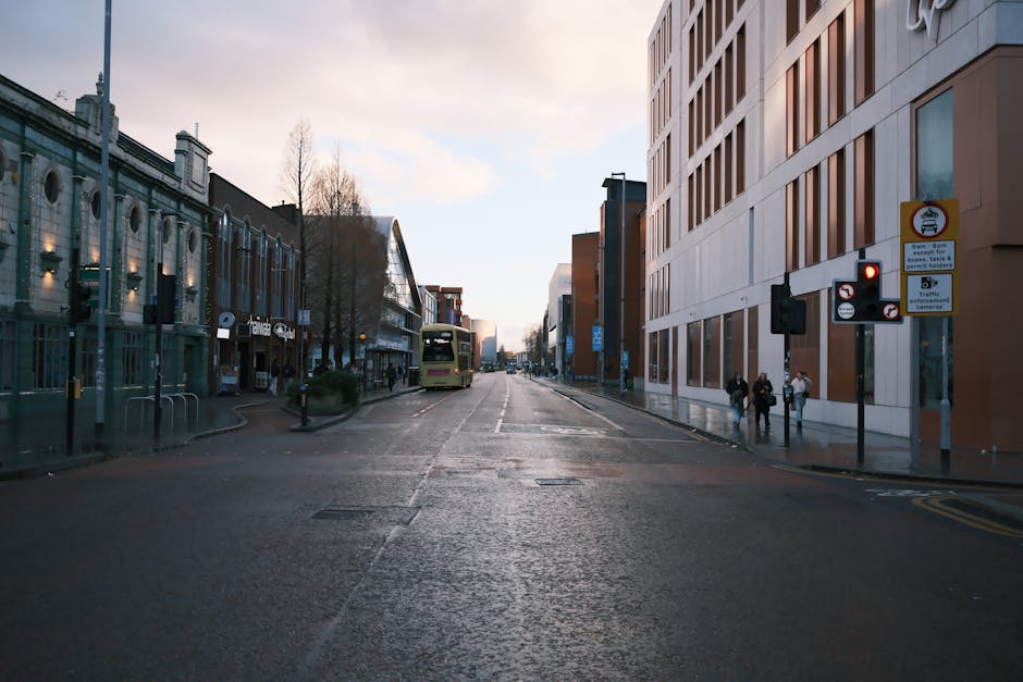 A wide urban street scene during daylight with a mix of commercial and modern office buildings on either side. On the left, a glass-fronted building houses shops with signage, while on the right, a contemporary multi-storey office block features large windows and a neutral facade. The street is relatively quiet, with a yellow bus visible in the distance and a few pedestrians walking on the pavement near the corner, some carrying bags. The road surface appears slightly wet, reflecting the overcast sky above. Traffic signals and street signs are mounted on poles at the corner, indicating parking restrictions and pedestrian crossings. In the foreground, the road is clear with no parked vehicles, creating space that could easily accommodate a small man with van for home relocation or furniture transport. The scene captures a typical city centre environment suitable for house removals, with surroundings conducive to loading and unloading goods as part of the moving process. Man with Van Preston, a professional removals service, could assist with such logistics during a move along this route, supporting efficient and careful packing and transport of household belongings.