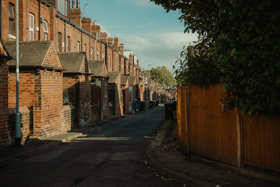 A narrow residential street in Preston showing a row of brick terraced houses with small front gardens and brick chimneys. The street is lined with black wheelie bins and a wooden fence on the right side, partially covered by green foliage. The pavement is in good condition, with some shadows cast by the buildings and trees. In the background, the street curves slightly, leading to more houses and trees beyond. The scene is captured during daylight, with a partly cloudy sky overhead, and the area appears quiet, suitable for house removals and furniture transport by [COMPANY_NAME], as part of home relocation and packing and moving services.