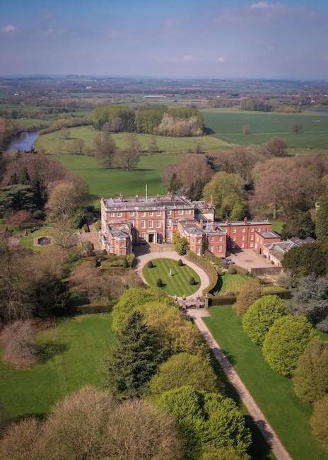 An aerial view of Fulwood Hall Estate showing a large, historic brick mansion surrounded by well-maintained gardens and lush green lawns. The estate is situated amid expansive rural landscape with fields, scattered trees, and a river visible in the distance under a partly cloudy sky. The image captures the estate’s driveway leading to the entrance, highlighting a tranquil setting ideal for house removals and furniture transport. This area is commonly associated with residential relocations, where Man with Van Preston provides professional packing and moving services to facilitate home relocations for large families. The scene emphasizes the estate’s grandeur and peaceful countryside environment, suitable for comprehensive moving logistics and house removal planning.
