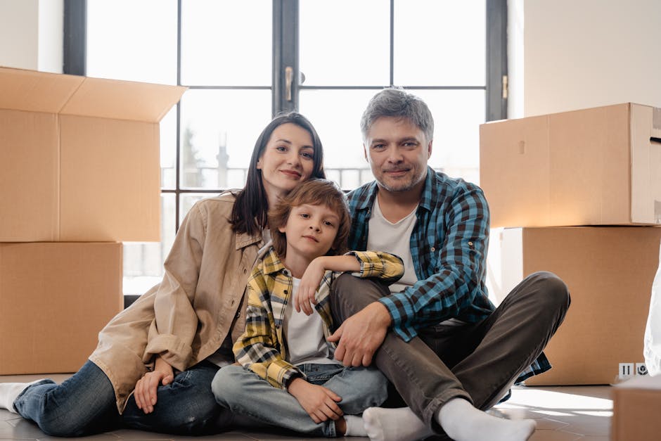 A family of three, comprising a woman, a man, and a young boy, sitting on the floor inside a home surrounded by packed cardboard boxes of various sizes, some sealed with packing tape and others open revealing contents. They are positioned near a large window with black framing, allowing natural daylight to illuminate the room. The woman, with shoulder-length dark hair and wearing a beige jacket, sits on the left, smiling gently. The young boy, with light brown hair and dressed in a plaid shirt, leans against the woman with his arm resting on her lap, looking at the camera. The man, with short gray hair and wearing a blue plaid shirt over a white T-shirt, sits on the right side, smiling slightly and holding onto one of the boxes. The background suggests a clean, organized space preparing for a home relocation, with cardboard boxes likely being ready for transport. This scene reflects the process of packing and moving, carried out by a professional removals service such as Man with Van Preston, illustrating the packing and loading stages of house removals in Fulwood, Preston.