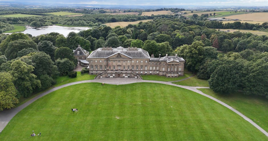 An aerial view of Fulwood Hall Estate showing a large, historic brick mansion surrounded by well-maintained gardens and lush green lawns. The estate is situated amid expansive rural landscape with fields, scattered trees, and a river visible in the distance under a partly cloudy sky. The image captures the estate’s driveway leading to the entrance, highlighting a tranquil setting ideal for house removals and furniture transport. This area is commonly associated with residential relocations, where Man with Van Preston provides professional packing and moving services to facilitate home relocations for large families. The scene emphasizes the estate’s grandeur and peaceful countryside environment, suitable for comprehensive moving logistics and house removal planning.