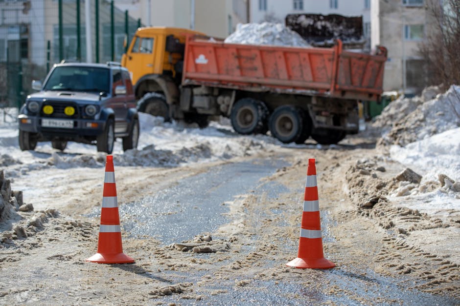 A construction or roadwork scene is visible, with a partially excavated dirt and gravel road marked off by two orange and white traffic cones in the foreground. Behind the cones, there are tire tracks and loose gravel scattered across the surface, indicating ongoing work. In the background, a large yellow and orange dump truck filled with construction debris and rubble is parked on a slightly elevated area. To the left, a dark-colored van with yellow headlights is parked near a fence, while further back, residential apartment buildings with multiple windows are visible. The area appears to be an urban setting, possibly in Preston, with snow present on the ground and surrounding areas. This image relates to the logistical challenges of home relocation and furniture transport, emphasizing the importance of planning for parking permits and access during moving services, as offered by Man with Van Preston.