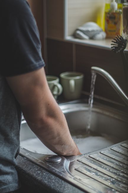 A person with their arm partially visible, standing next to a kitchen sink with running water, appears to be cleaning or preparing items for a house move. The scene shows the stainless steel sink with