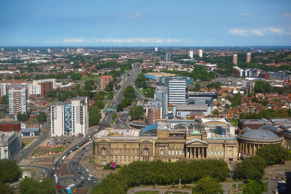 An aerial view of Preston cityscape showing a mix of modern and historic buildings, with roads and streets busy with moving vehicles, including a large stone building with classical architecture in the foreground. Residential and commercial properties, green spaces, and high-rise structures are visible in the distance under a partly cloudy sky, providing context for house removals and furniture transport activities often associated with home relocation services, as provided by Man with Van Preston.