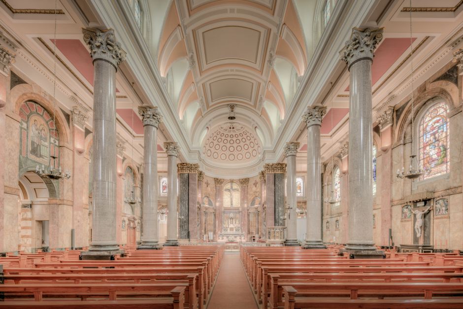 Interior view of a large church or cathedral with classical architectural features, including tall grey marble columns with ornate capitals supporting a high, decorated ceiling with intricate moldings and circular designs. The space is illuminated by natural light coming through stained glass windows on both sides, which depict religious scenes and figures. Wooden pews lined in rows face an altar area at the far end, which appears to be used for worship or ceremonial purposes. The interior walls are decorated with religious statues and artwork, with some areas featuring alcoves, wall-mounted candleholders, and frescoes. The floor is wooden, and moving materials such as cardboard boxes, wrapped furniture, or packing supplies are not visible in this image, indicating a focus on the historic architecture rather than a removal or relocation process. The overall scene emphasizes the spaciousness and detailed craftsmanship typical of religious buildings, with natural light enhancing the warm tones of the wooden furnishings and the pinkish hues of the walls.