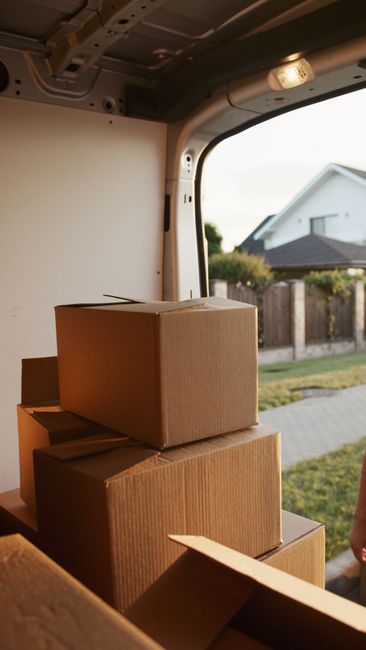 Inside the rear of a van during a home relocation, several brown cardboard boxes of varying sizes are stacked on a flat surface near the open rear door. The boxes are sealed with packing tape, and some are partially open, revealing packing materials. The van's interior features a metal ceiling with mounted fixtures and wiring visible. Outside the open door, a residential street with a white house, a wooden fence, and a paved footpath is visible in the background, illuminated by natural daylight. This scene reflects the packing and loading process involved in furniture transport and house removals, with Man with Van Preston providing professional removals services.
