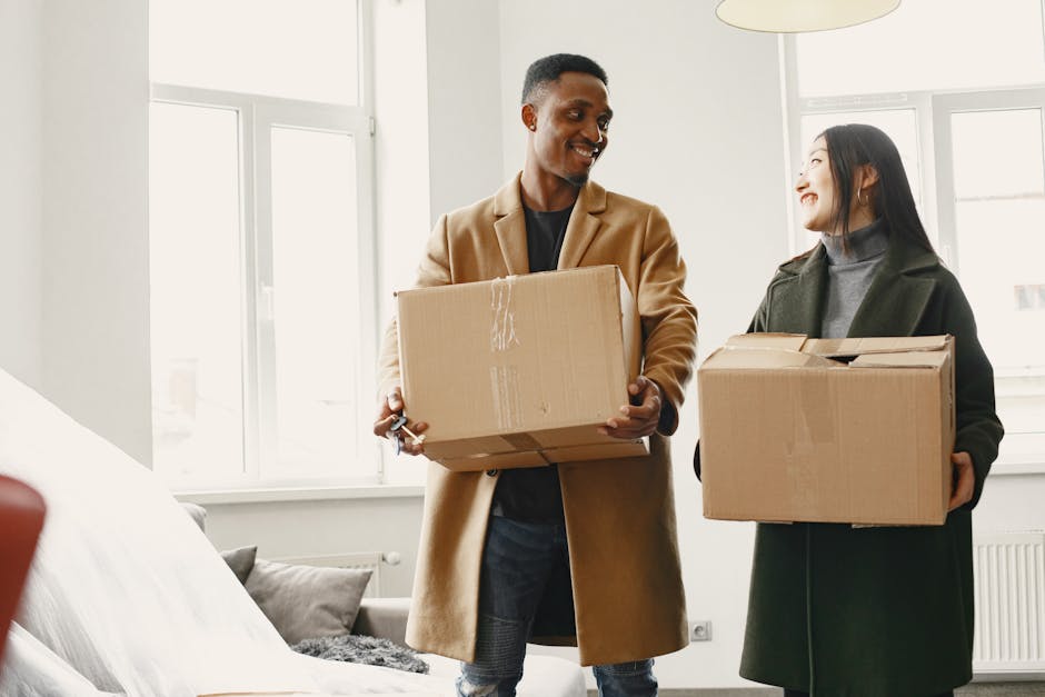 A man with dark skin and short hair, wearing a tan coat and black shirt, is holding a cardboard box while smiling at a woman with pale skin and long dark hair, dressed in a dark coat and grey turtleneck, who is also holding a cardboard box inside a well-lit residential room. The background features white walls with large windows allowing natural light to fill the space. A beige sofa with cushions can be seen in the corner. This scene depicts a home relocation process, where both individuals are engaged in packing or unpacking as part of a furniture transport or moving procedure, coordinated by Man with Van Preston, a professional removals service. The image captures the moment of collaboration during a short-notice student move in Preston, focusing on the handling of boxes and the interior environment, highlighting the logistics involved in packing and moving within a domestic setting.