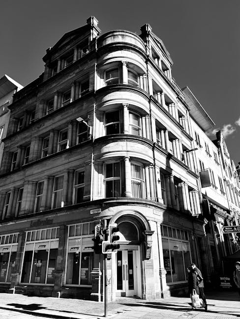 A black-and-white photograph of a multi-storey building with ornate architectural details, including curved bay windows and decorative stonework, situated on a city street. The building's ground floor features large windows and an arched entrance, with a traffic light and pedestrian crossing in front. A person is standing near the curb with a suitcase, while another individual appears to be waiting further along the pavement. Inside the building, visible through open doorways, are packing materials such as cardboard boxes and plastic wrapping, indicating a home relocation process. Some furniture or appliances may be partially visible through the windows, and the overall scene suggests the external phase of furniture transport or packing and moving activities undertaken by a professional removals service, such as Man with Van Preston, in preparation for a house move or student relocation during term-time.