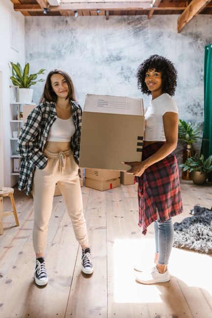 A man with dark skin and short hair, wearing a tan coat and black shirt, is holding a cardboard box while smiling at a woman with pale skin and long dark hair, dressed in a dark coat and grey turtleneck, who is also holding a cardboard box inside a well-lit residential room. The background features white walls with large windows allowing natural light to fill the space. A beige sofa with cushions can be seen in the corner. This scene depicts a home relocation process, where both individuals are engaged in packing or unpacking as part of a furniture transport or moving procedure, coordinated by Man with Van Preston, a professional removals service. The image captures the moment of collaboration during a short-notice student move in Preston, focusing on the handling of boxes and the interior environment, highlighting the logistics involved in packing and moving within a domestic setting.