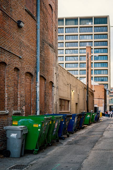 A row of large wheelie bins in green, blue, and grey, lined up along a narrow urban street next to a brick building with a concrete wall. The bins are positioned on the pavement, with some partially open, and are used for waste disposal, likely for household or commercial rubbish. Behind the bins, a grey drainpipe runs vertically down the brick wall, and a small window is visible on the concrete section of the building. In the background, there is a modern multi-storey office building with numerous windows, suggesting a city environment. The scene appears to be part of the load or collection process related to home or business rubbish removal, relevant to house removal or moving logistics, with subtle associations to the need for waste clearance during relocations. Man with Van Preston, a professional removals company, often handles such logistics as part of their comprehensive service in Preston.