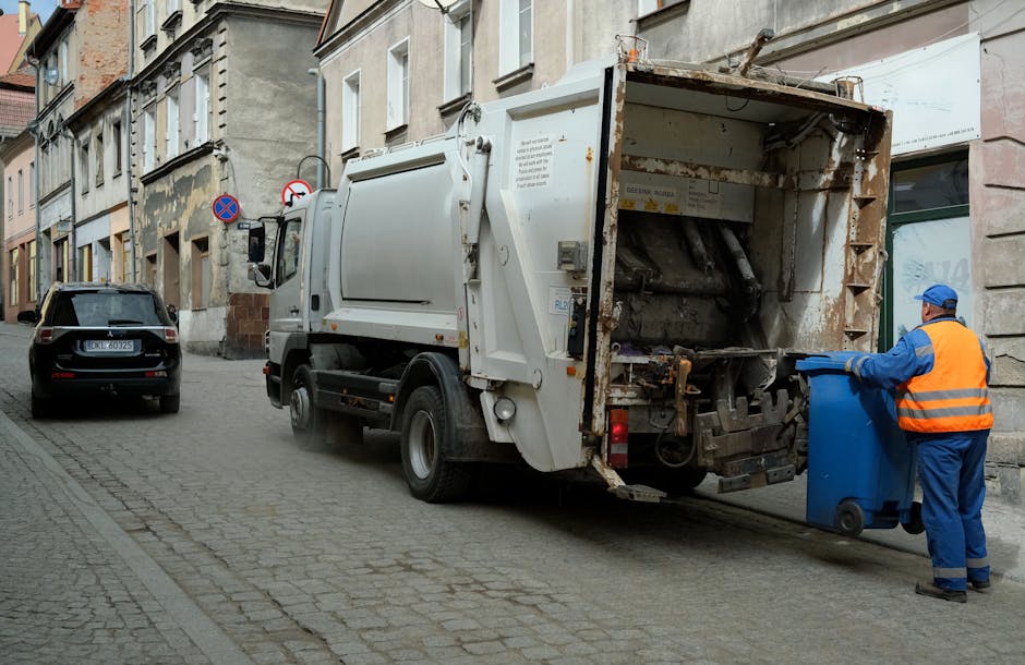 A refuse collection truck parked on a narrow cobblestone street in Preston, with a worker in a high-visibility orange vest and blue uniform operating a blue wheeled bin directly in front of the truck. The truck has a large, open rear loading area, with visible metal components and some rust, and is positioned near a residential building with a stone facade. Several cardboard boxes and plastic-wrapped household items are inside the truck's compartment. The worker is in the process of loading or unloading waste or bulky waste items, which may include furniture or packaging materials, supporting the home relocation or removals process. A black car is parked further along the street, and the scene is illuminated by natural daylight, with typical town street features in the background.