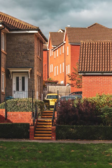 A residential street scene showing a terraced home with a front garden and a staircase leading to a white front door. The house has a grey brick exterior and a tiled roof. Behind and beside it are taller red-brick houses with multiple windows and gabled tiled roofs. Several parked cars, including a yellow vehicle, are visible along the pavement near the properties. The garden is bordered by low brick walls and lush green hedges, with some bushes and grass in the foreground. Overhead, a cloudy sky is seen, casting diffuse lighting over the scene. This image captures the typical environment where house removals and furniture transports might occur, with a focus on the proximity of the buildings and the external features of the properties, illustrating the logistical considerations involved in home relocation services provided by Man with Van Preston.