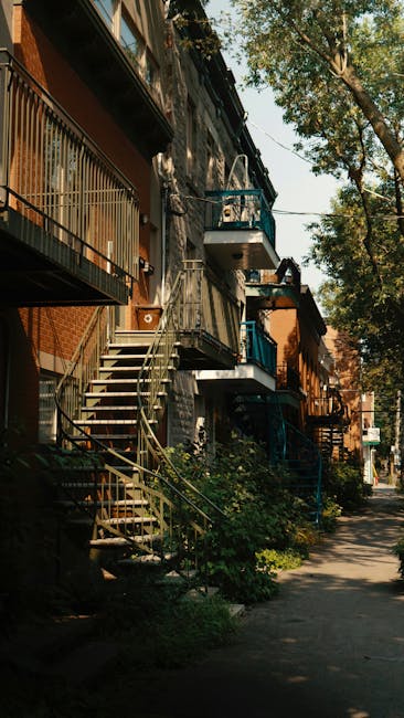 Exterior view of a row of traditional terraced homes with narrow staircases leading from ground level to small balconies, situated along a tree-lined street in Preston. The buildings are made of brick with some painted facades, and the staircases feature metal railings; some stairs are curved, with visible steps and railings. Sunlight filters through the branches of tall trees, casting shadows on the pavement and building facades. The sidewalk in front includes plants and shrubs, and the street appears quiet, with no visible pedestrians or vehicles. This setting reflects typical urban housing with narrow stairs suited for home relocation and furniture transport. The image is taken during daytime with natural light emphasizing the architecture and environment. Man with Van Preston provides removals and moving services in such residential areas, assisting with packing and loading processes for house relocations involving such narrow stairwells and terraced homes.