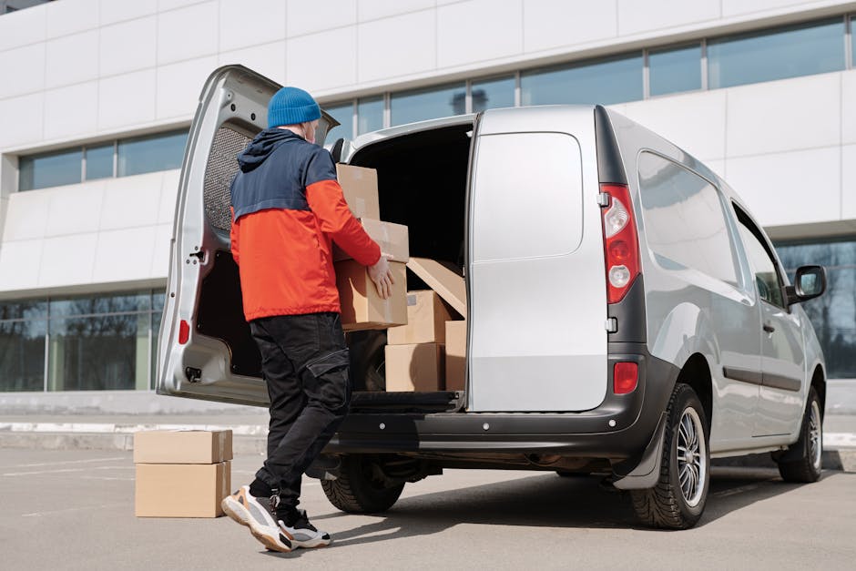 Two men are engaged in a home relocation process outside a residential property, utilizing a hand truck to carry a large cardboard box marked with fragile handling warnings. One man is lifting the box
